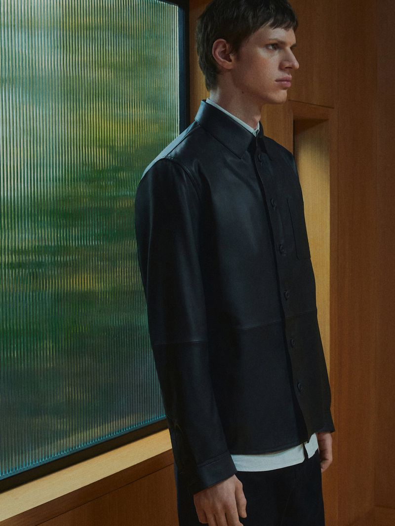 Portrait of a fair-skinned adult male in a black leather jacket and black pants, standing indoors near a window, medium shot, eye-level angle.