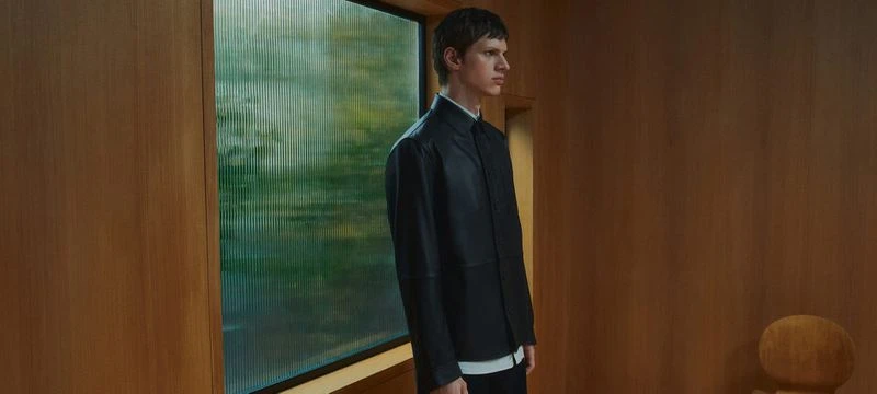Adult male in black jacket stands indoors near window with textured glass, soft lighting, against wooden wall.