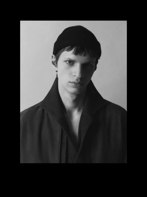 Monochrome portrait of an adult male model wearing a black beanie, dark overcoat with a thick collar, and dark V-neck sweater, against a grey backdrop.