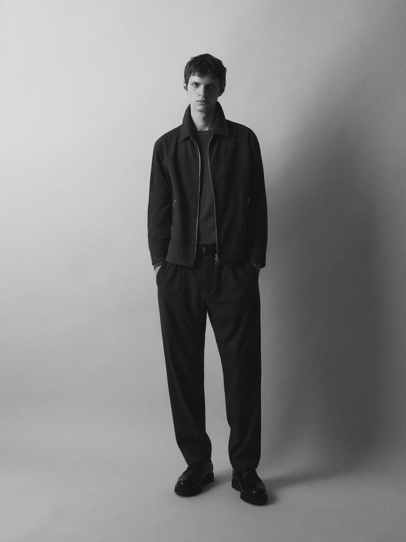 Monochrome portrait of a young adult man in a dark jacket and trousers against a muted grey background, shot in a studio setting.