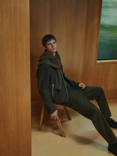 An adult male model sits on a folding stool in an earth-toned jacket, scarf, and pants, with a wood paneled wall in the background.