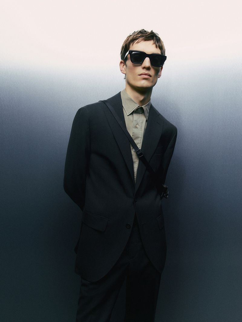 Man in black suit and sunglasses stands against a gradient gray background, with a shoulder bag and a neutral expression.