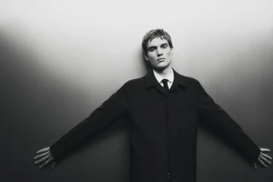 Man wearing a black coat and tie leaning against a seamless grey studio wall for Massimo Dutti