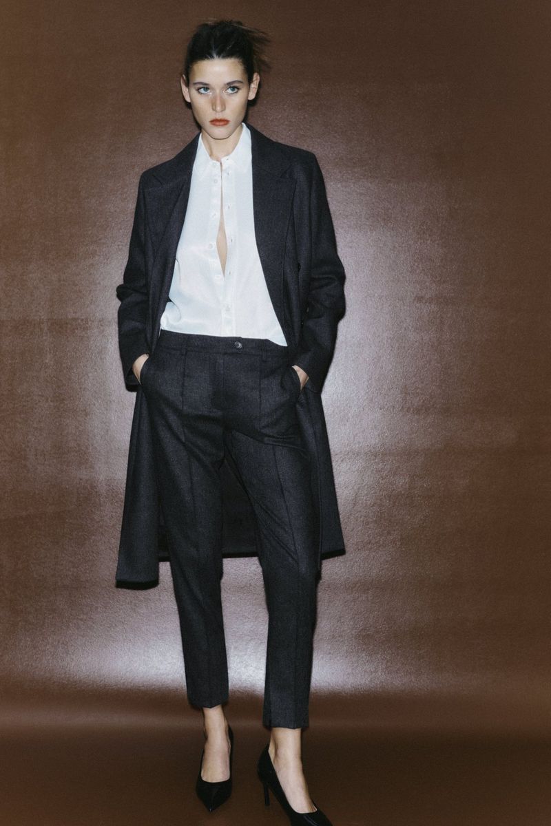 Fashionable brunette woman wearing a dark coat, white blouse, dark pants and black heels posing against a solid brown backdrop.