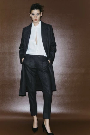 Fashionable brunette woman wearing a dark coat, white blouse, dark pants and black heels posing against a solid brown backdrop.