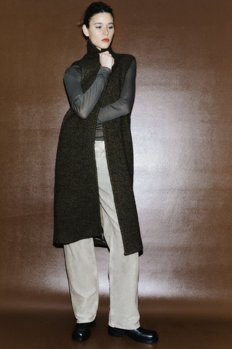 A woman models a long, dark-green sweater vest with light-beige trousers and black flats against a brown backdrop in a studio shot.