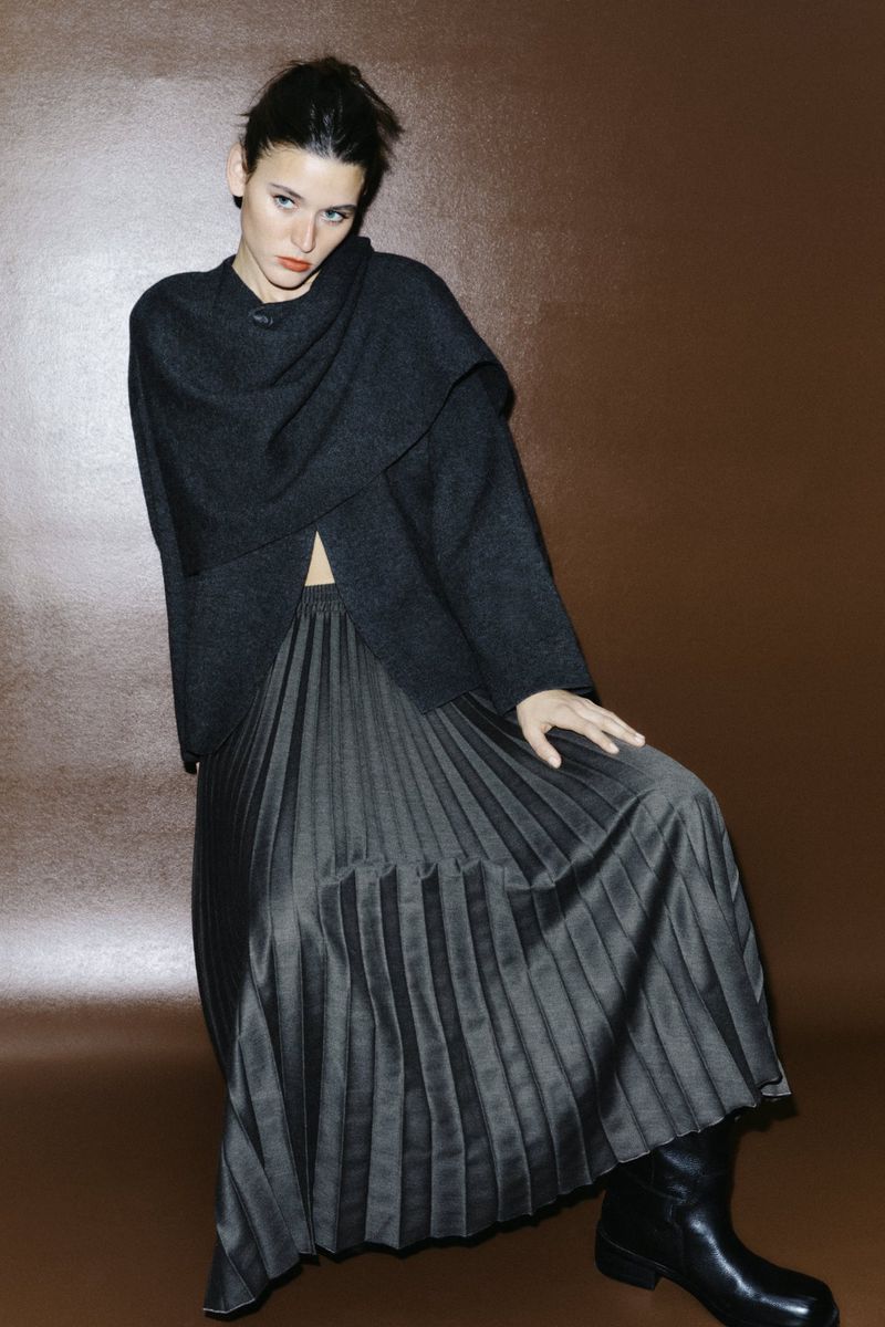 Fashionable woman in dark sweater and pleated midi skirt poses in studio, set against a simple brown backdrop for modern, edgy style.