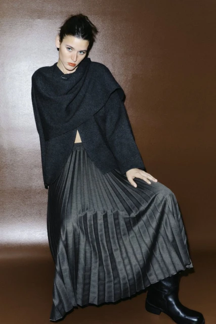 Fashionable woman in dark sweater and pleated midi skirt poses in studio, set against a simple brown backdrop for modern, edgy style.