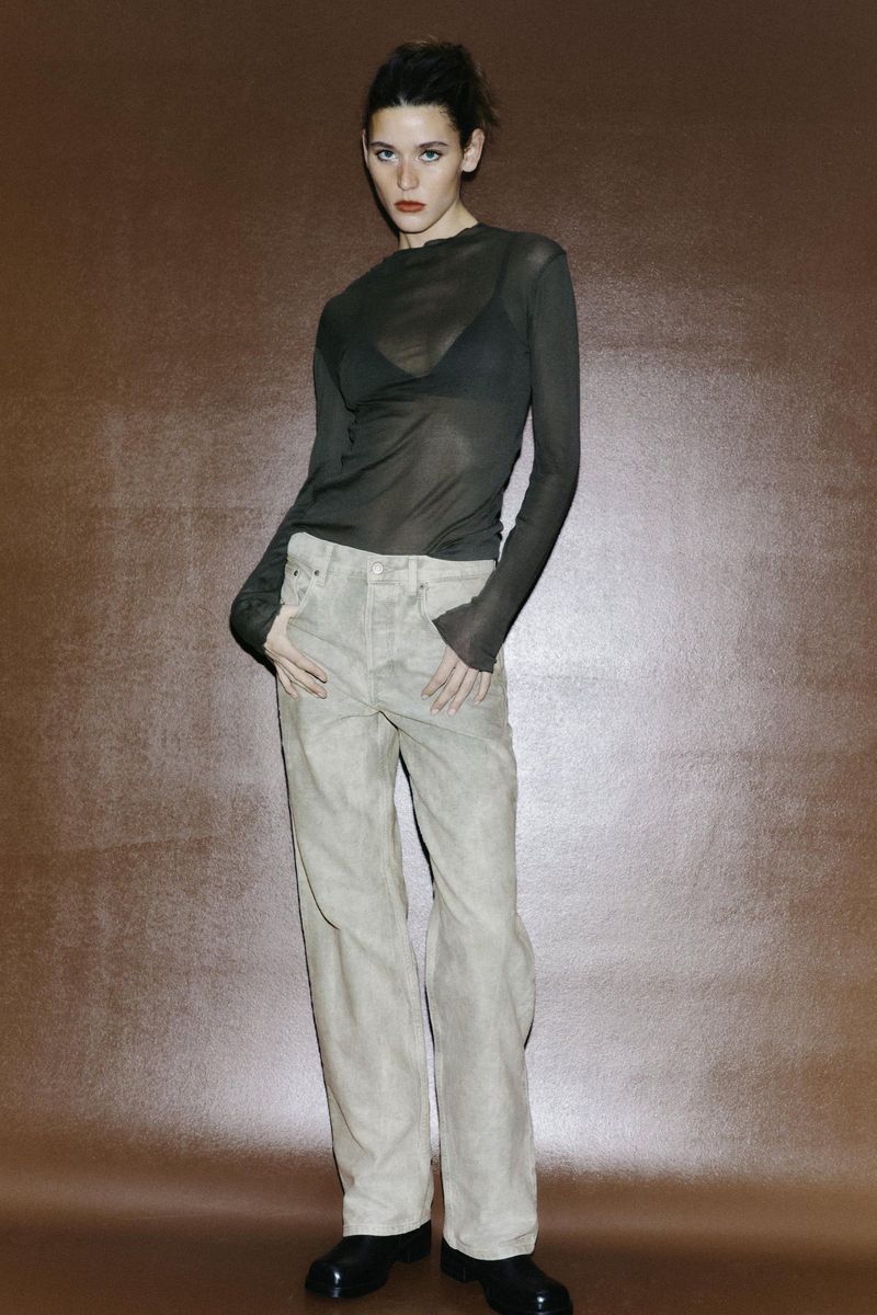 Full-length studio shot of a young adult woman posing in a sheer dark green long-sleeve top, beige pants, and black patent leather shoes.
