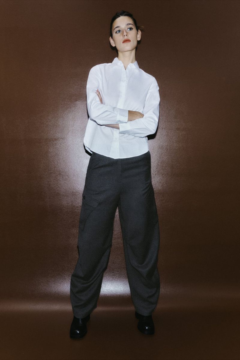 Full-length studio shot of a female model in a white button-down shirt and oversized dark pants against a plain brown backdrop.