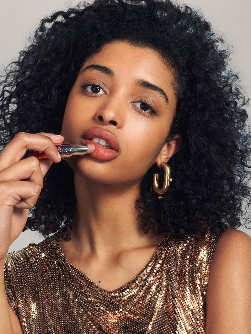 Close-up of a beautiful black woman applying nude lipstick and wearing gold hoop earrings and a gold, metallic v-neck top.