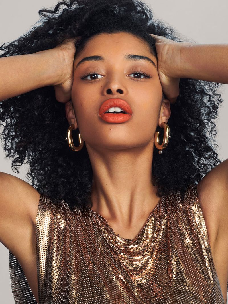 Close-up portrait of an African American woman with curly hair wearing gold earrings and a metallic gold top against a white backdrop.