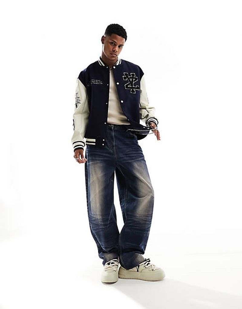 Full-length shot of a young adult male in a varsity jacket and baggy jeans, standing against a white background in a studio.
