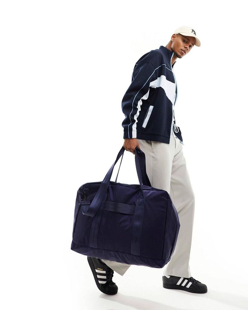 A dark-skinned male model carries a navy duffel bag and poses in a studio, wearing a navy track jacket, cream trousers, and black sneakers.