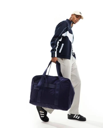 A dark-skinned male model carries a navy duffel bag and poses in a studio, wearing a navy track jacket, cream trousers, and black sneakers.
