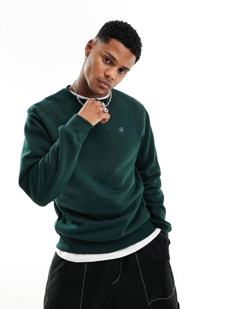 A young adult male poses in front of a white backdrop wearing a dark green sweater with black pants and silver chain necklace.