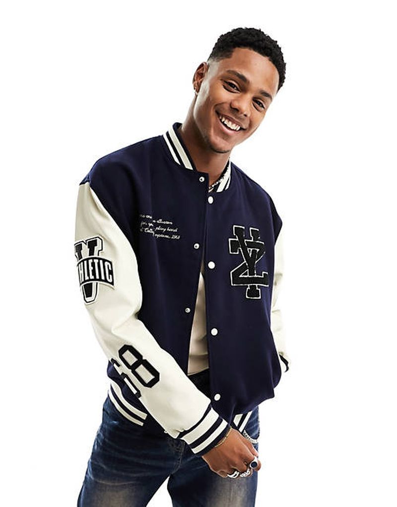 A young adult male smiles, wearing a navy and white varsity jacket with lettered patches, posing against a white backdrop in a studio shot.