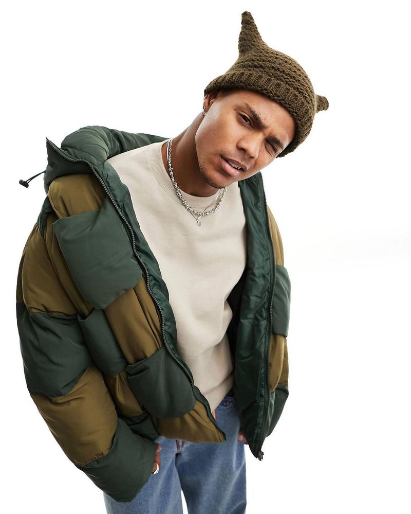 Young adult man in a quilted puffer jacket and knit elf hat poses in a studio against a white backdrop with a coy expression.