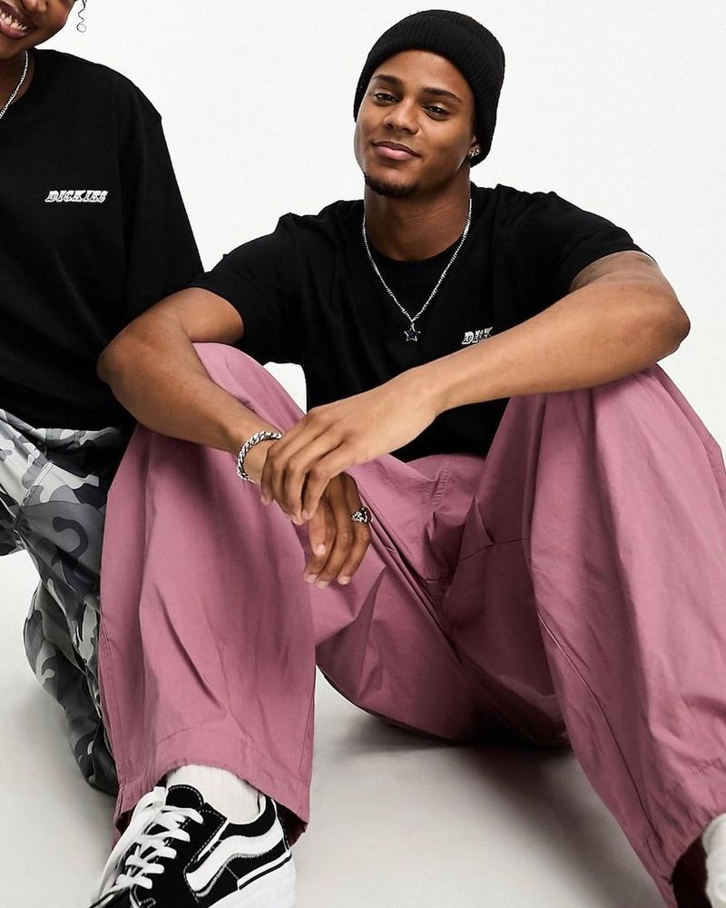 Two people wearing black shirts and pink pants sit indoors. One person wears a black beanie and a silver star necklace, and sneakers.