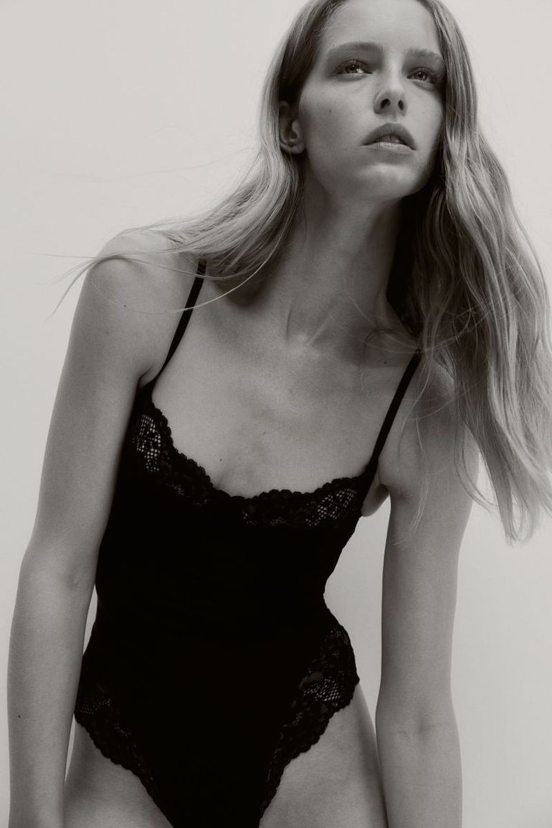 A woman with long blonde hair poses in a photo studio wearing a black, lace-trimmed bodysuit against a neutral backdrop.