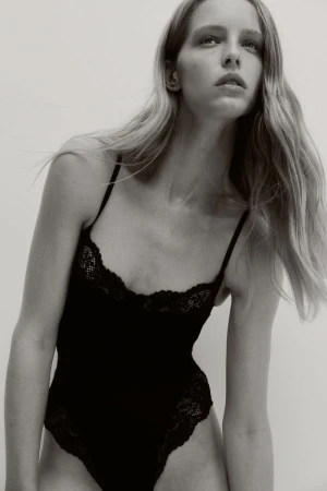 A woman with long blonde hair poses in a photo studio wearing a black, lace-trimmed bodysuit against a neutral backdrop.