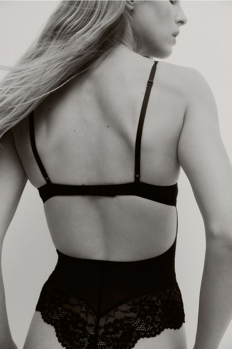 Back view of an adult female model with long blonde hair wearing black lace lingerie against a plain light background in a photo studio.