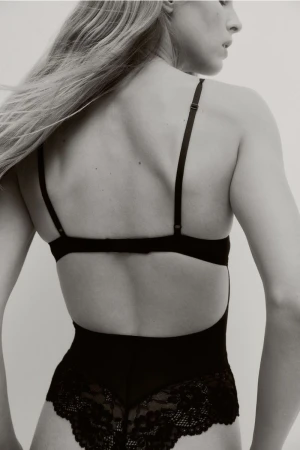 Back view of an adult female model with long blonde hair wearing black lace lingerie against a plain light background in a photo studio.