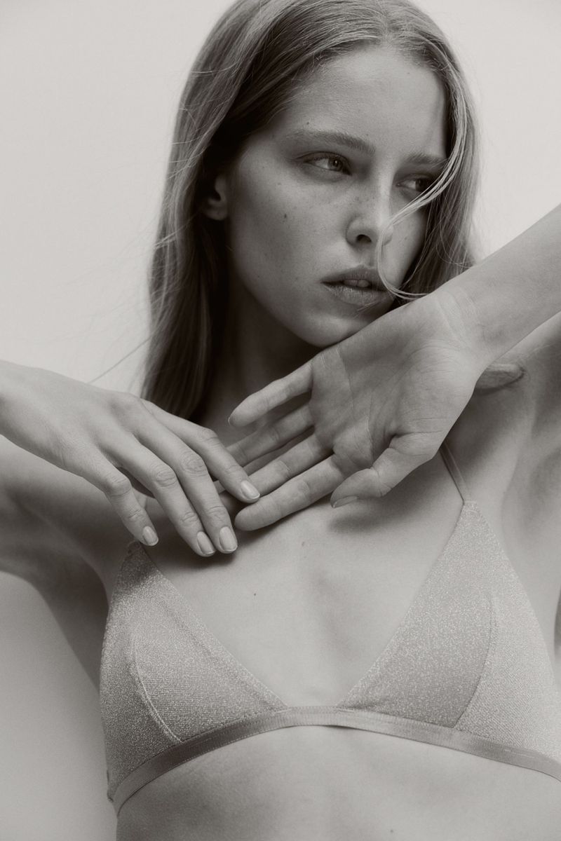 Black-and-white portrait of a young adult female model in a triangle bra with hands raised, posing in front of a plain white background.