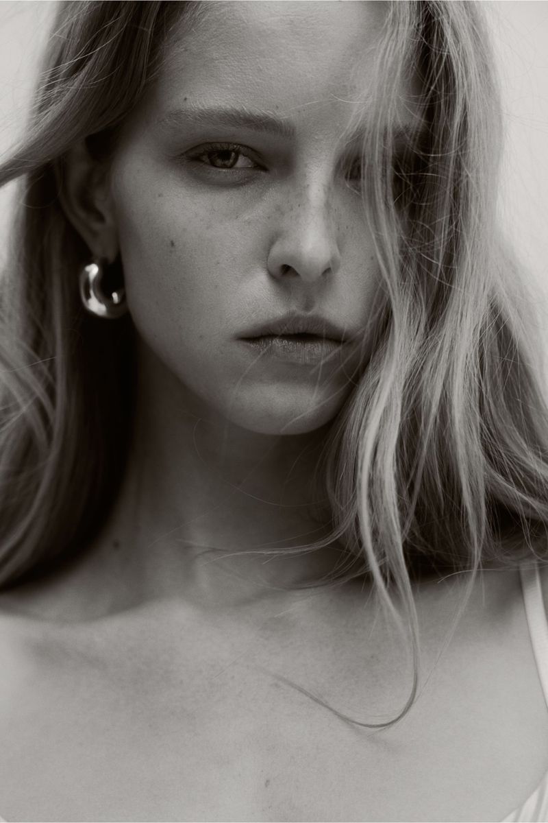 Close-up portrait of a blonde adult woman with a serious expression wearing hoop earrings against a neutral, light-toned background.