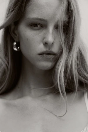 Close-up portrait of a blonde adult woman with a serious expression wearing hoop earrings against a neutral, light-toned background.
