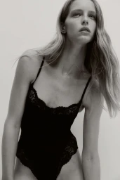 Model Abby Champion wearing a black lace bodysuit against a plain light background for an H&M lingerie campaign.