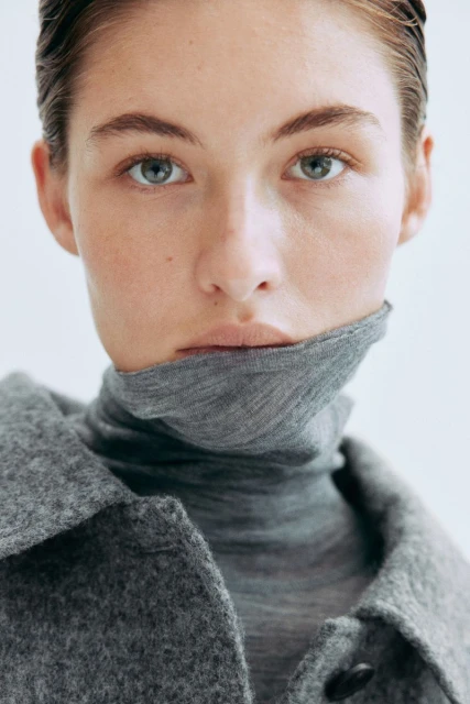 Close-up portrait of a fair-skinned woman with wet-look hair and grey-green eyes, wearing a grey turtleneck sweater and wool coat against a white backdrop.