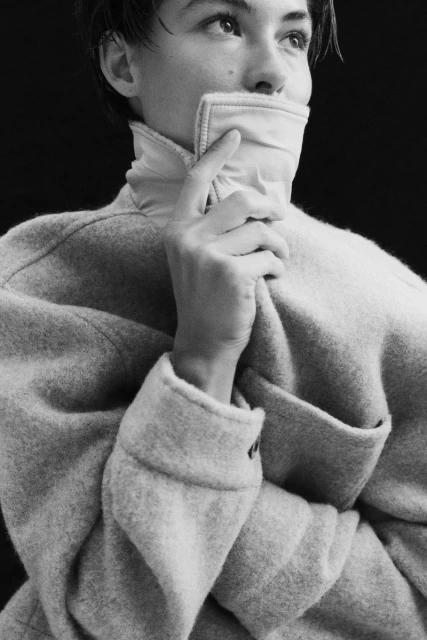 Monochrome portrait of a person in a wool coat with a high collar, holding the collar with one hand, looking up and to the right.