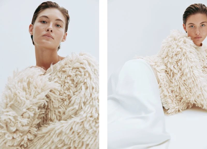 Fashion shot of a young adult woman in an off-white, shaggy sweater and white pants in a studio setting, looking at the camera.