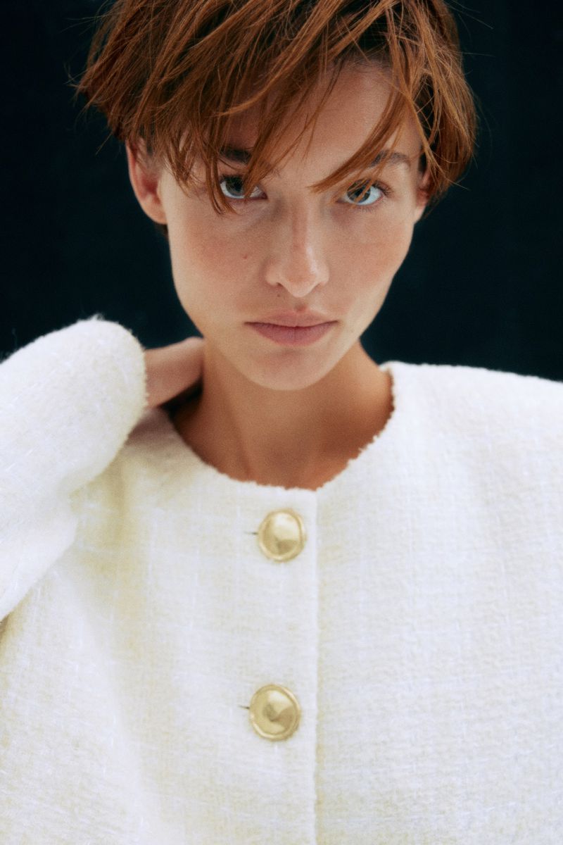 Close-up portrait of an adult woman with short auburn hair wearing a white bouclé jacket with gold buttons against a dark background.
