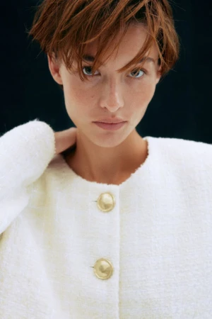 Close-up portrait of an adult woman with short auburn hair wearing a white bouclé jacket with gold buttons against a dark background.