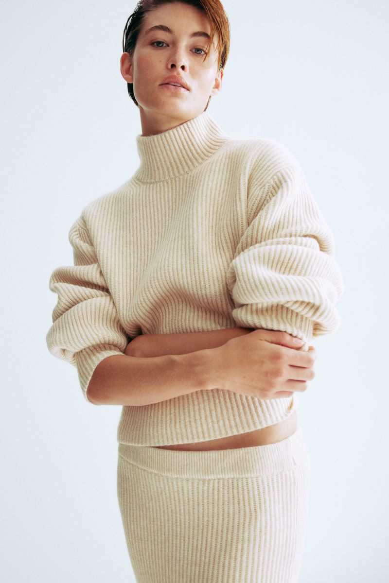 Indoor studio shot of a young adult female wearing a ribbed beige sweater with a high neckline and a matching skirt, posing.