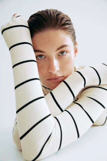 Close-up studio shot of a young, light-skinned woman with blue eyes, wearing a striped long-sleeve shirt, posing with her arms.