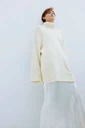 Model wearing a chunky cream turtleneck sweater and white skirt against a white studio background.