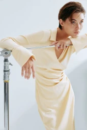 Model wearing a pale yellow collared dress leaning against a metal studio C-stand against a white backdrop.