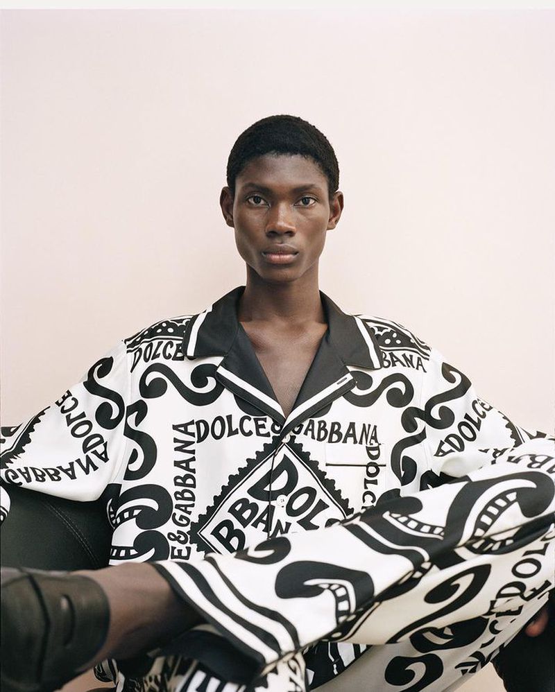Portrait of a Black male model in patterned Dolce & Gabbana pajamas sitting in a chair against a light pink studio background.