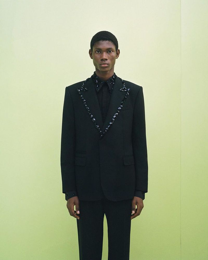 Man in black suit with embellished trim stands against a muted yellow backdrop, offering a minimalist, formal fashion portrait.