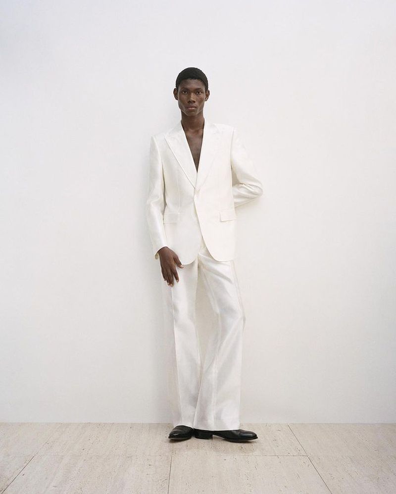 An adult African American male model poses in a white suit against a white wall in a studio. Fashion shot, full length.
