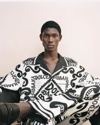 Black man seated wearing a matching black and white patterned silk shirt and trousers by Dolce&Gabbana for Farfetch.