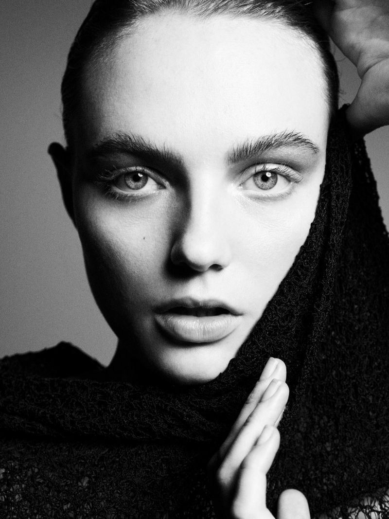 Monochrome portrait of a woman with dark hair and light eyes, posing with hands near her face and wearing a textured black garment.