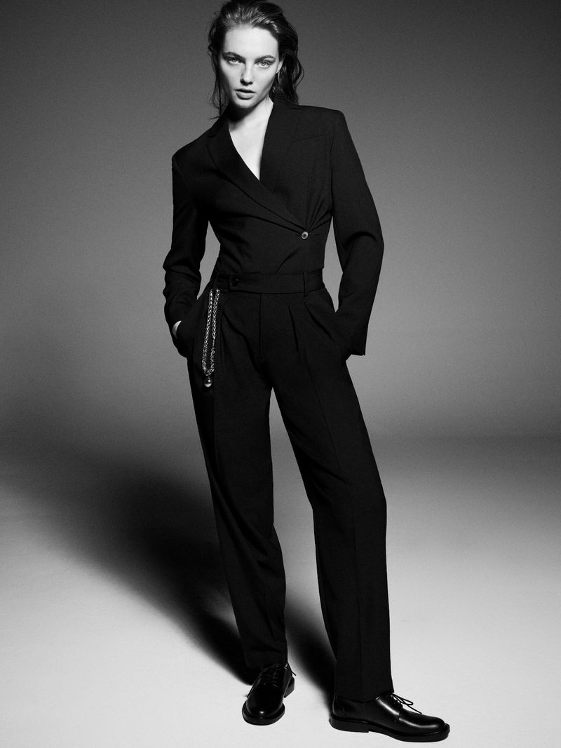 A stylish studio shot of a woman in a black jumpsuit and dress shoes, complemented by a delicate chain detail against a grey backdrop.