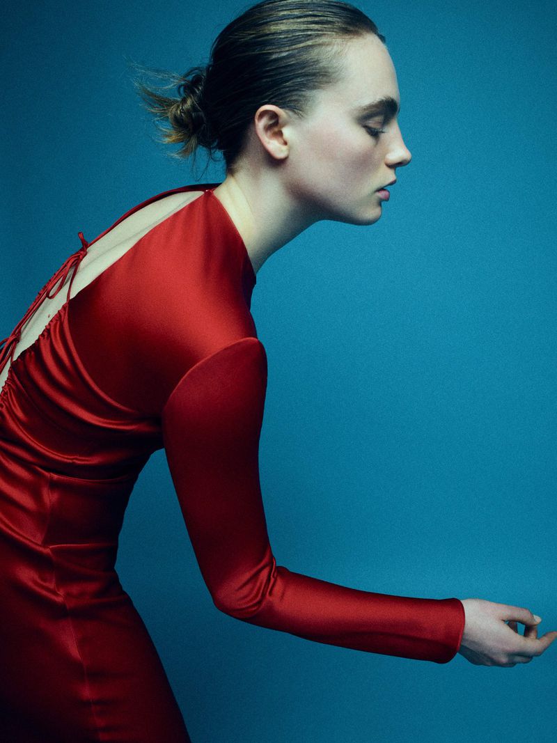 Profile view of a woman with her hair in a bun wearing a red, long-sleeved dress against a blue background in a studio setting.