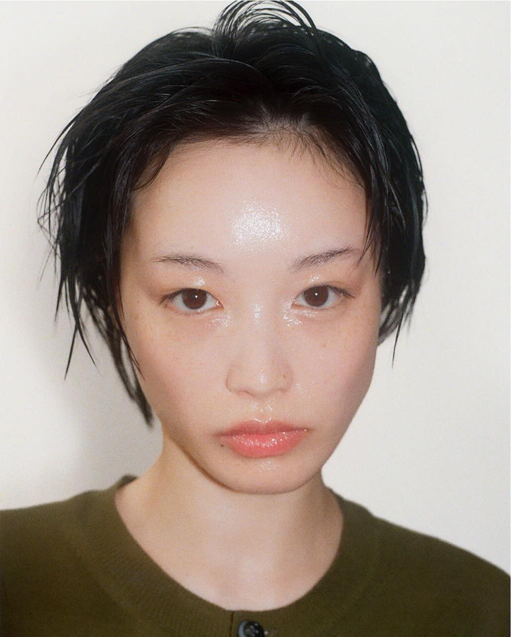 Close-up portrait of a young, fair-skinned East Asian woman with short black hair and an olive green sweater against a white background.