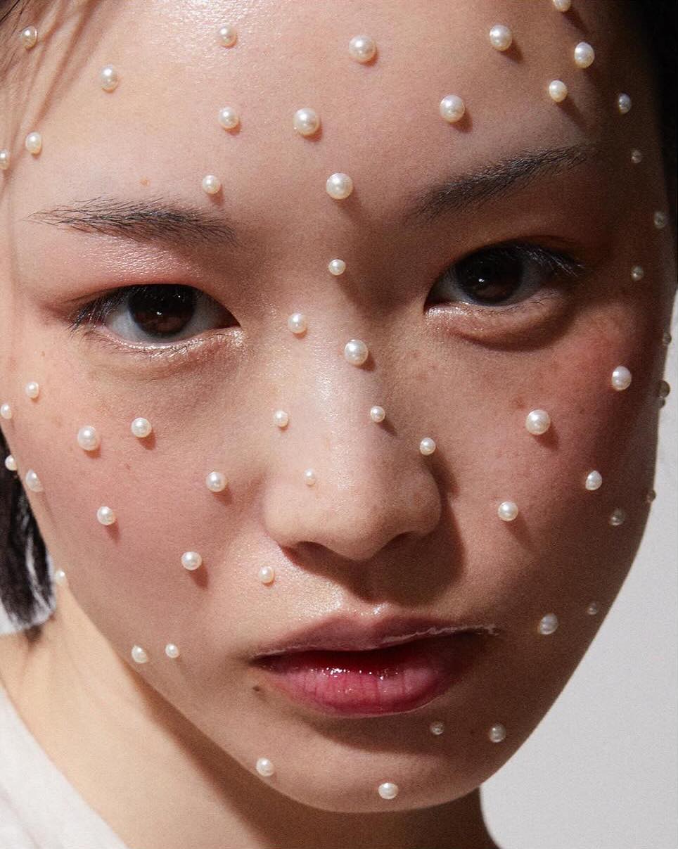 Close-up portrait of a young Asian female model with fair skin adorned with numerous small, round pearls on her face in a beauty context.