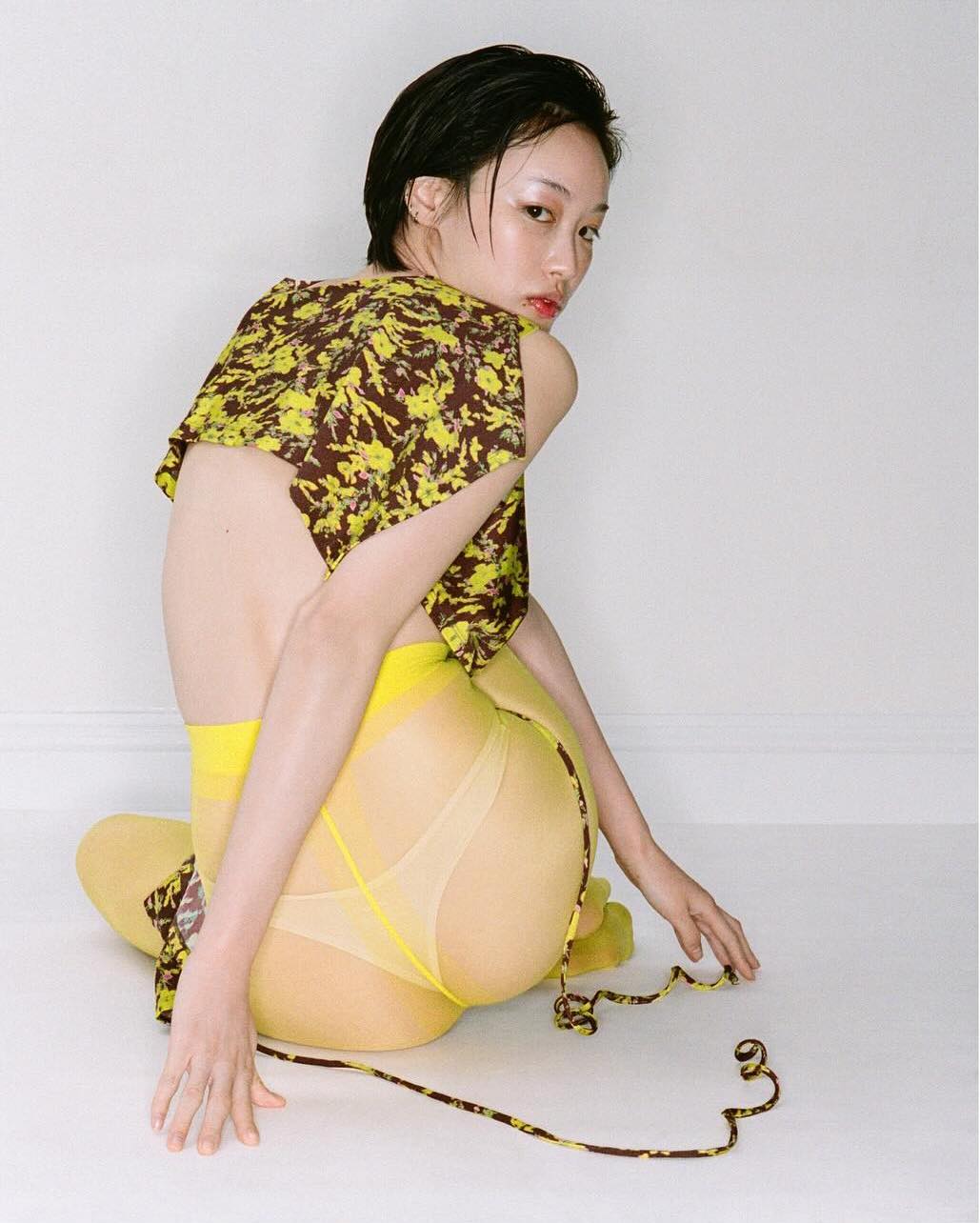 An adult female sits in a photo studio wearing a crop top, nylon hosiery, and decorative straps, posing against a white wall.